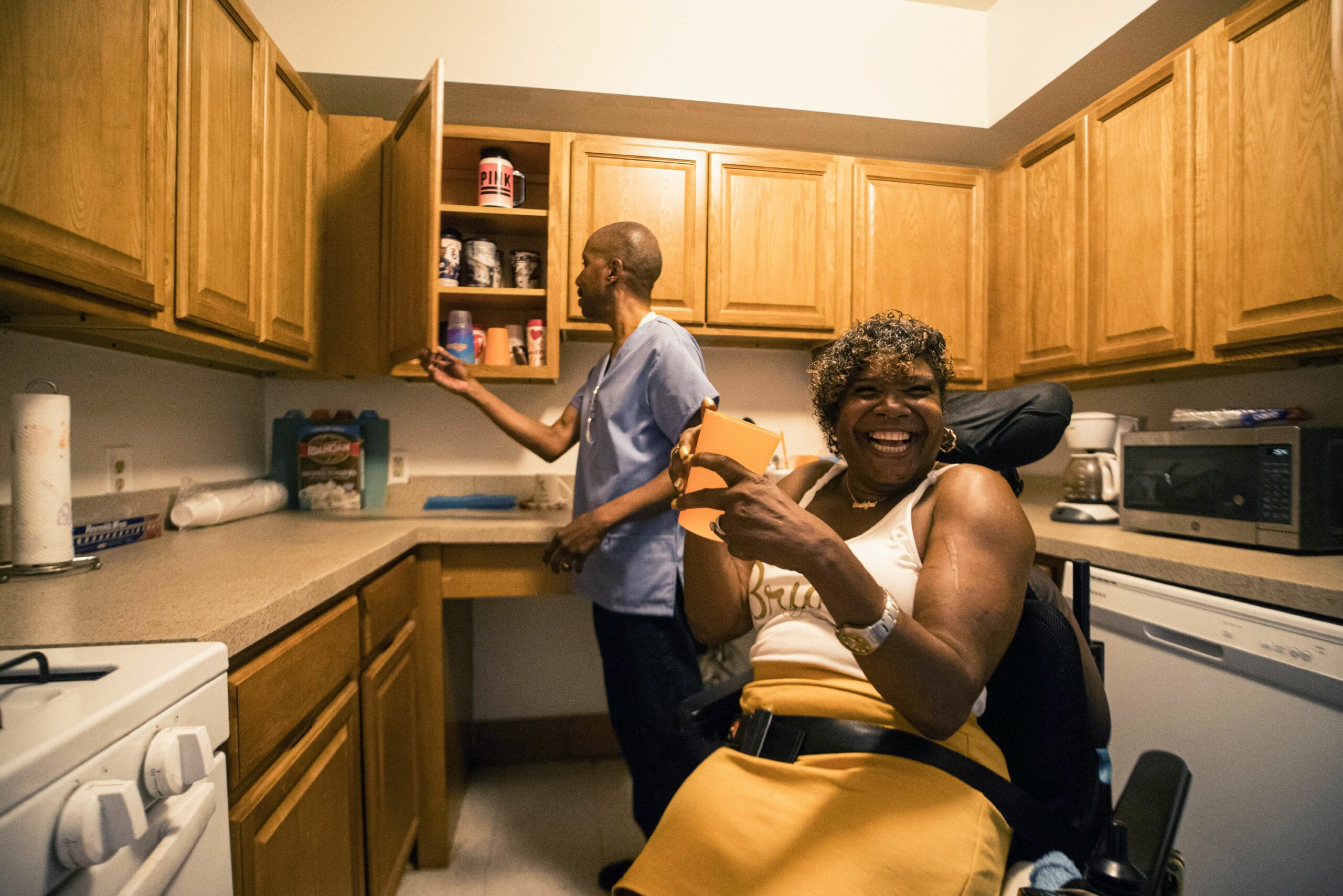 Woman in wheelchair smiling at camera while caregiver looks in cabinet